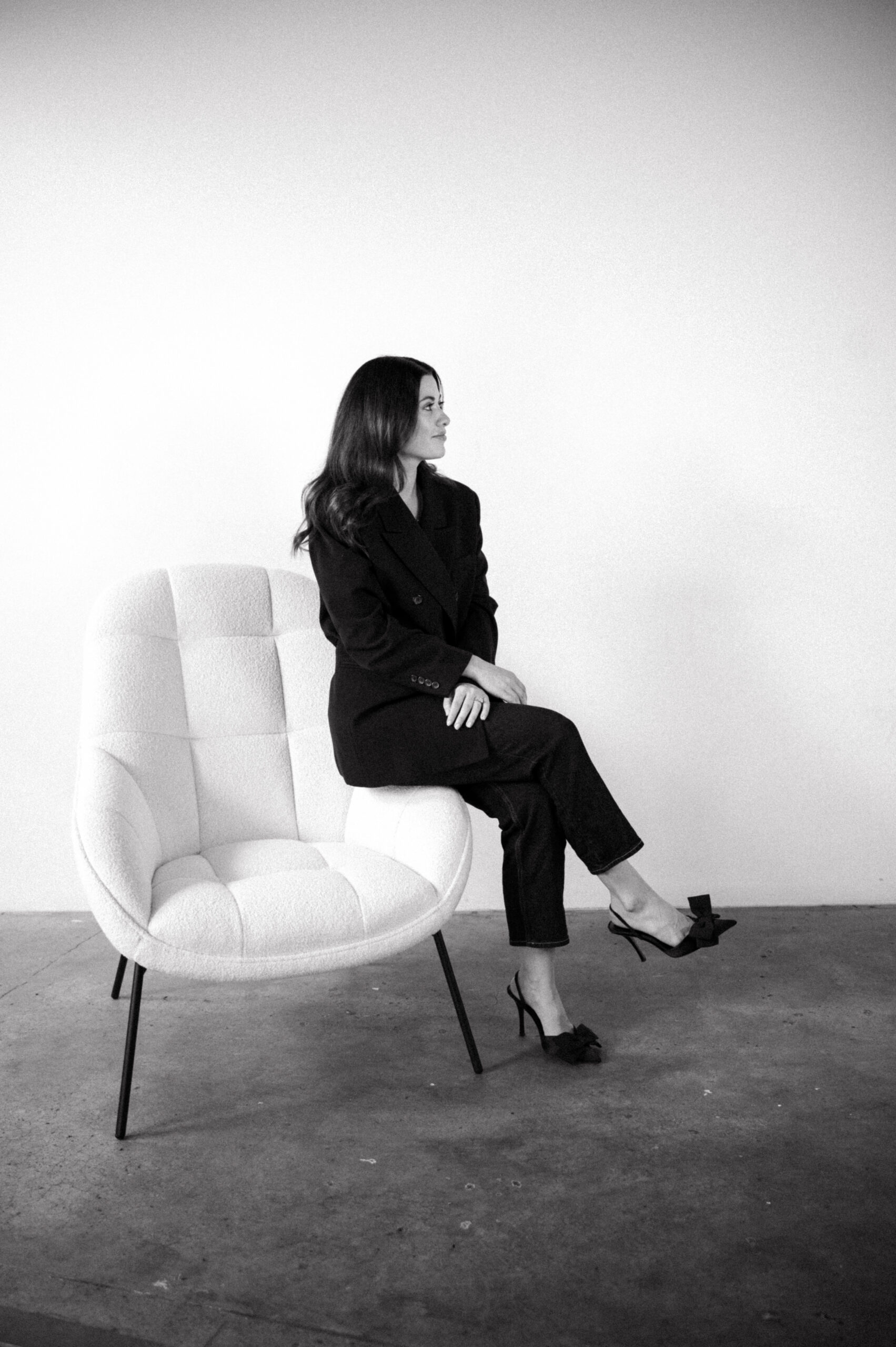 Black and white headshot of Daniella Boughen, Founder & Digital Director of Digible. Daniella is perched on the side of a white occasional chair with her legs crossed.